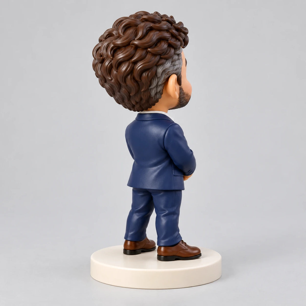 Dreiviertelansicht von hinten einer Chibi-Männerfigur mit gewelltem braun-grauem Haar, marineblauem Anzug und braunen Schuhen auf einem elfenbeinfarbenen runden Sockel.